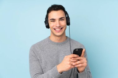 Kafkasyalı yakışıklı adam mavi arka planda izole edilmiş. Cep telefonuyla müzik dinliyor ve ön tarafa bakıyor.