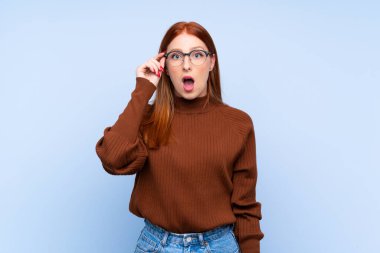 Genç kızıl saçlı kadın gözlüklü ve şaşırmış bir şekilde mavi arka planda.