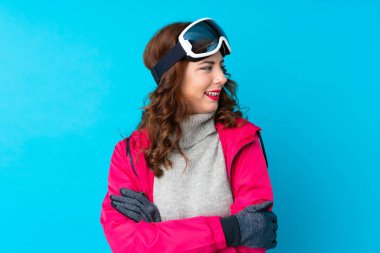 Snowboard gözlüklü kayakçı kadın mavi duvarın üzerinden yan tarafa bakıyor.