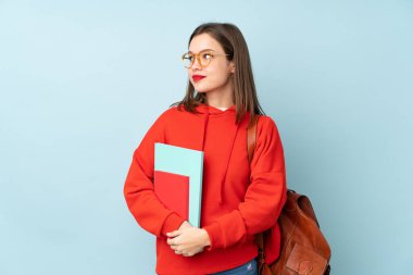 Mavi arka planda izole edilmiş kitapları tutan öğrenci kız ayakta duruyor ve yan tarafa bakıyor.