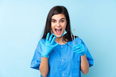 Genç kadın diş hekimi, mavi arka plan üzerinde sürpriz yüz ifadesiyle aletler tutuyor.