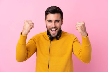 Kulaklıkları olan genç yakışıklı bir adam tecrit edilmiş pembe arka planda zaferi kutluyor.