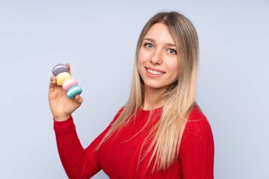 Soyutlanmış mavi arka planda genç sarışın kadın elinde renkli Fransız kurabiyeleri ve bol bol gülümsüyor.