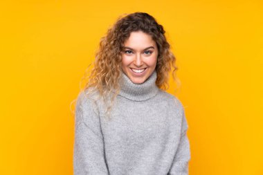 Sarışın, kıvırcık saçlı, balıkçı yaka kazak giyen, sarı arka planda izole edilmiş, gülen genç bir kadın.