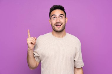 Young handsome man over isolated purple background pointing up and surprised