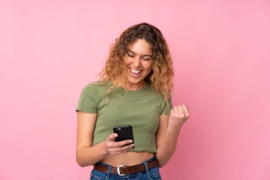 Pembe arka planda izole edilmiş kıvırcık saçlı sarışın genç bir kadın ve telefon zafer pozisyonunda.