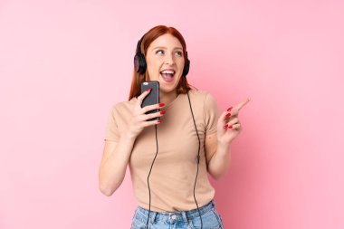 Genç kızıl saçlı kadın pembe arka planda, kulaklıklı ve şarkı söyleyen cep telefonunu kullanıyor.