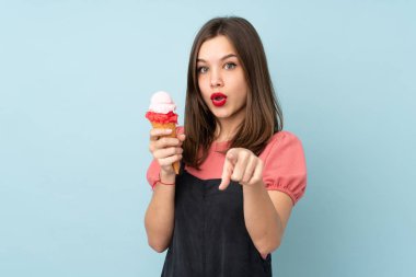Kornet dondurması tutan genç kız mavi arka planda izole edilmiş şaşırmış ve ön tarafı işaret ediyor.