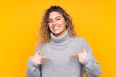 Sarışın, kıvırcık saçlı, balıkçı yaka kazak giyen, sarı arka planda soyutlanmış gururlu ve kendini beğenmiş bir kadın.