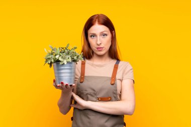 Genç, kızıl saçlı bahçıvan kadın, sarı arka planda bir bitki tutuyor, kollarını kavuşturuyor.