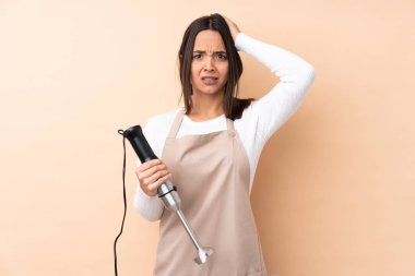 Genç esmer kız el blenderi kullanıyor izole edilmiş arka planda hayal kırıklığına uğramış ve ellerini başının üstüne koyuyor.