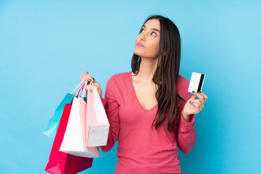 Genç esmer kadın izole edilmiş mavi arka planda elinde alışveriş çantaları ve bir kredi kartıyla düşünüyor.