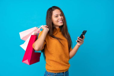 Genç Brezilyalı kız mavi arka planda, elinde alışveriş çantaları ve cep telefonuyla bir arkadaşına mesaj yazıyor.