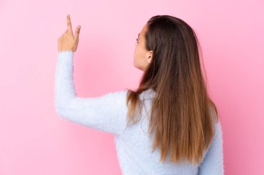 İzole edilmiş pembe arka planı olan mavi kazaklı genç kız işaret parmağıyla işaret ediyor.