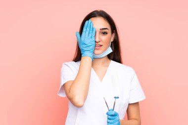 Pembe arka planda izole edilmiş aletleri tutan dişçi kadın bir gözü elle kapatıyor.