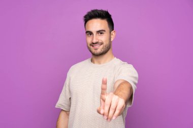 İzole edilmiş mor arka planda genç yakışıklı bir adam parmağını kaldırıyor ve gösteriyor.