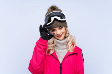 Snowboard gözlüklü genç kız izole edilmiş mavi arka planda parmaklarıyla iyi bir işaret gösteriyor.