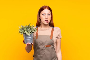 Genç, kızıl saçlı bahçıvan kadın, sarı arka planda bir bitki tutuyor. Şüpheleri var ve kafası karışık.