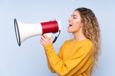 Mavi arka planda izole edilmiş kıvırcık saçlı genç sarışın kadın megafondan bağırıyor.