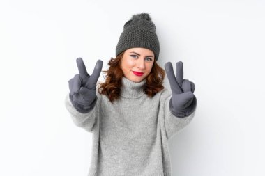 Kışlık şapkalı genç Rus kadın izole beyaz arka planda gülümsüyor ve zafer işareti gösteriyor.