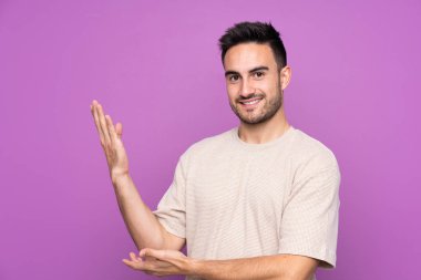 Soyutlanmış mor arka planda genç yakışıklı adam, davet edilmek için ellerini yana uzatıyor.