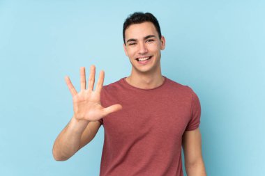 Beyaz tenli yakışıklı adam mavi arka planda izole edilmiş, parmaklarıyla beşi sayıyor.