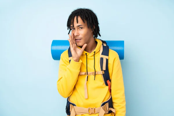 Young mountaineer african american man with a big backpack isolated on a blue background whispering something