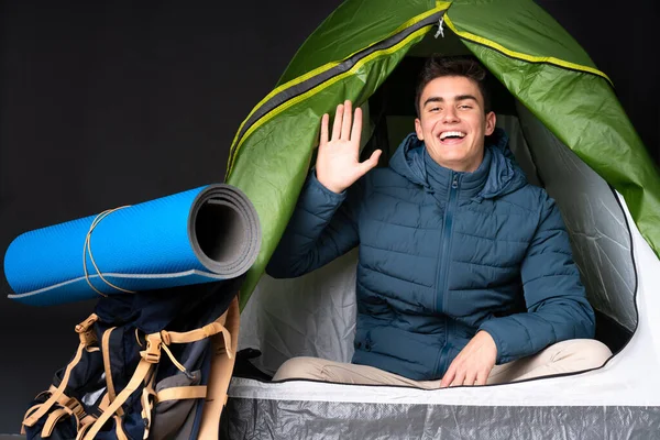 Teenager caucasian man inside a camping green tent isolated on black background saluting with hand with happy expression