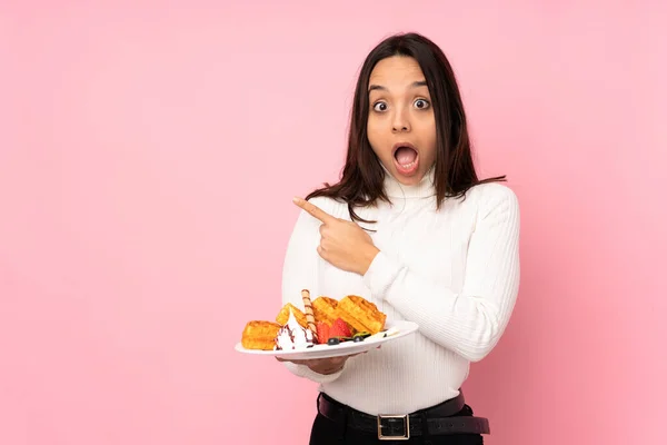 Young Brunette Woman Holding Waffles Isolated Pink Background Surprised Pointing — Stock Photo, Image