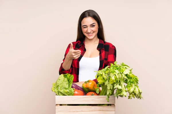 Farmer with freshly picked vegetables in a box isolated on beige background points finger at you