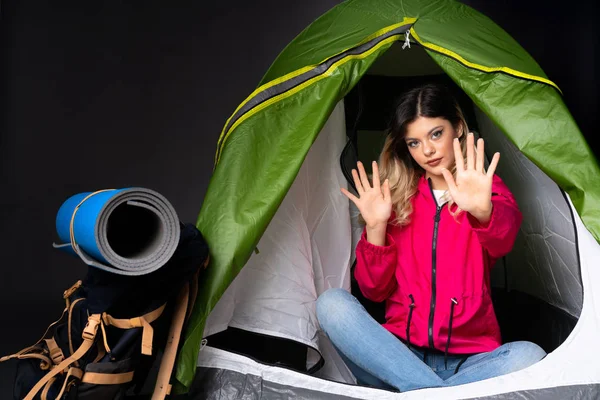 Teenager girl inside a camping green tent isolated on black background nervous stretching hands to the front