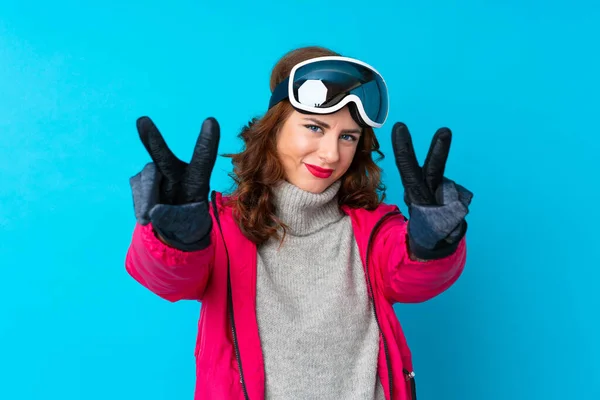 Skier woman with snowboarding glasses over isolated blue wall smiling and showing victory sign