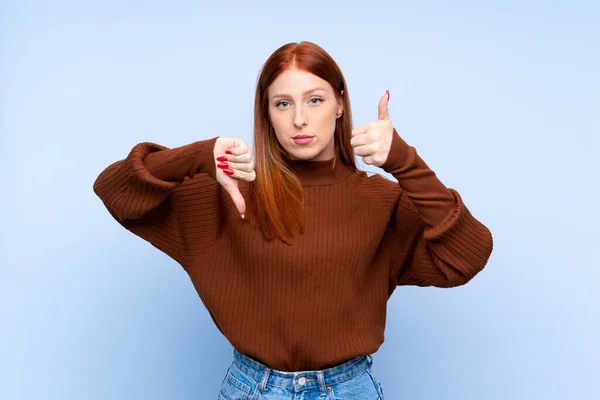 Young redhead woman over isolated blue background making good-bad sign. Undecided between yes or not