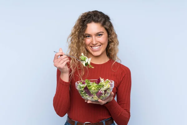 Jovem loira com cabelo encaracolado segurando uma salada sobre parede isolada — Fotografia de Stock