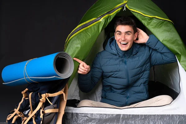 Teenager caucasian man inside a camping green tent isolated on black background surprised and pointing finger to the side