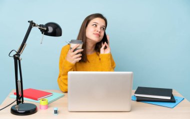 Öğrenci kız, mavi arka planda izole bir şekilde evinde ders çalışıyor. Elinde kahve ve cep telefonu var.