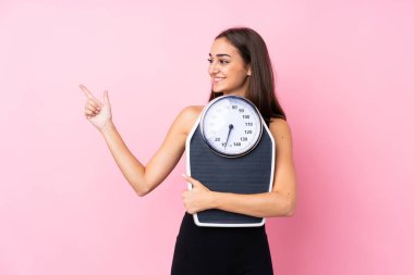 Tartı makinesi olan güzel bir genç kız, izole edilmiş pembe arka plan, tartı makinesi ve işaret eden bir tarafı var.