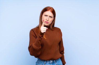 Genç kızıl saçlı kadın, izole edilmiş mavi arka planda hayal kırıklığına uğramış ve ön tarafı işaret ediyor.