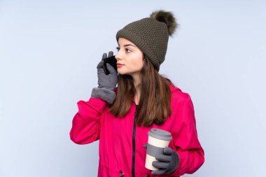 Kışlık şapkalı genç kız mavi arka planda izole edilmiş. Elinde kahve ve cep telefonu var.