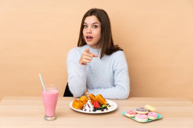 Bej arka planda izole edilmiş waffle yiyen genç kız şaşırmış ve ön tarafı işaret ediyor.