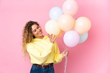 Kıvırcık saçlı genç sarışın kadın pembe arka planda izole edilmiş bir sürü balon yakalıyor.