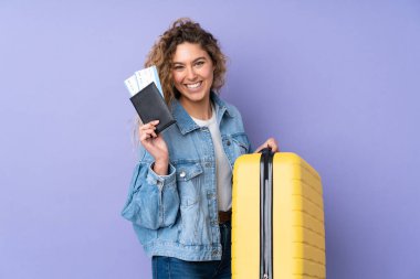 Kıvırcık saçlı genç bir kadın, mor arka planda izole edilmiş, bavul ve pasaportla tatilde.