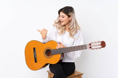 İzole edilmiş beyaz arka planda gitarı olan genç bir kız bir ürünü sunmak için kenarı işaret ediyor.