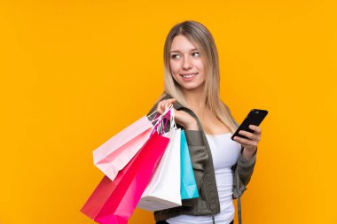 Sarışın, izole edilmiş sarı arka planda genç bir kadın. Elinde alışveriş çantaları ve cep telefonu var.
