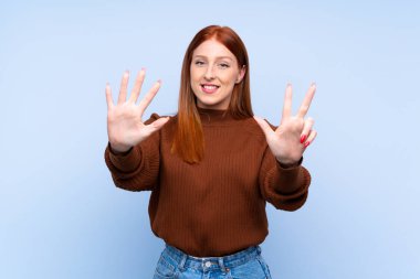 Genç kızıl saçlı kadın, izole edilmiş mavi arka planda sekiz parmaklı sayıyor.