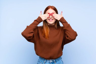 İzole edilmiş mavi arka planı olan genç kızıl saçlı kadın elleriyle gözlerini kapatıyor.