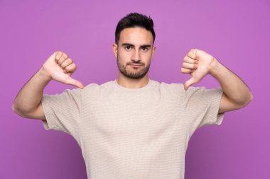 İzole edilmiş mor arka planda genç yakışıklı adam baş parmağını aşağı gösteriyor.