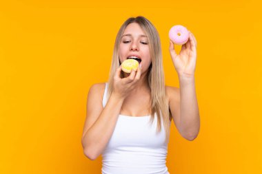 Soyutlanmış mavi arka planda genç sarışın bir kadın donut yiyor.