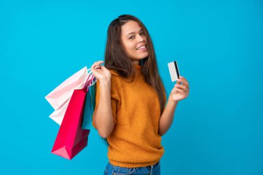 Genç Brezilyalı kız izole edilmiş mavi arka planda elinde alışveriş çantaları ve bir kredi kartı tutuyor.