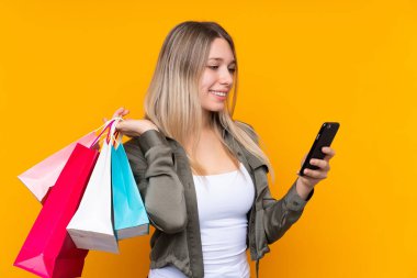 Sarışın, izole edilmiş, sarı arka planı olan genç bir kadın alışveriş torbaları tutuyor ve bir arkadaşına cep telefonuyla bir mesaj yazıyor.